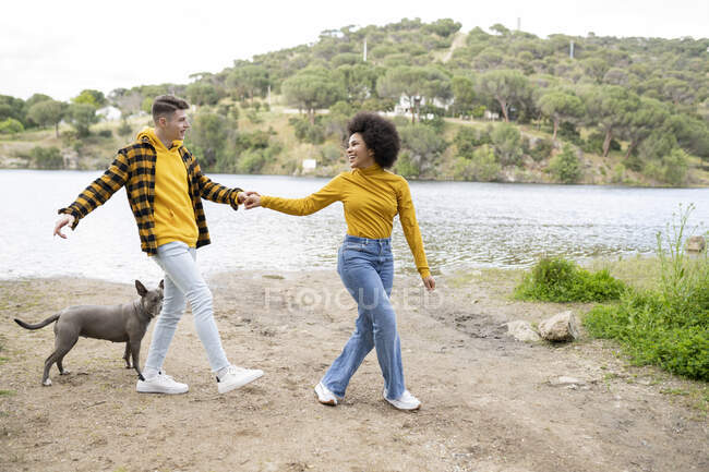 Corpo inteiro encantado multiétnico jovem e mulher de mãos dadas e olhando um para o outro enquanto caminhava com o cão na costa do rio na natureza — Fotografia de Stock
