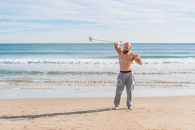 Volle Länge des erwachsenen sportlichen Mannes, der beim Aufwärmen an der sonnigen Küste wegschaut und dabei um seinen Körper herumgeht, mit Hantelscheibe am Band — Stockfoto