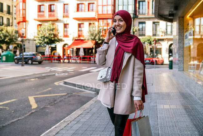 Mulher muçulmana alegre no hijab e com sacos de compras andando ao longo da rua e falando no smartphone — Fotografia de Stock