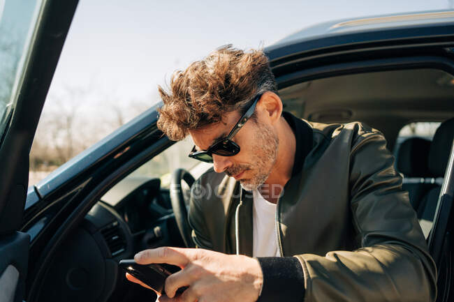 Серйозний чоловік в сонцезахисних окулярах переглядає мобільний телефон, сидячи в автомобілі в сонячний день — стокове фото