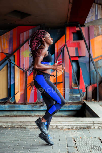 Вид сбоку на мускулистую афроамериканку в спортивной одежде, прыгающую высоко в воздухе во время тренировки возле стены современного здания на городской улице — стоковое фото