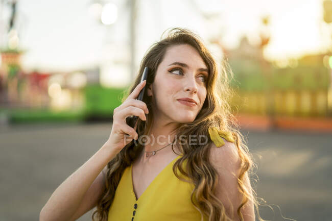 Contenu femelle parlant sur téléphone portable tout en se tenant dans le parc d'attractions en soirée en été — Photo de stock