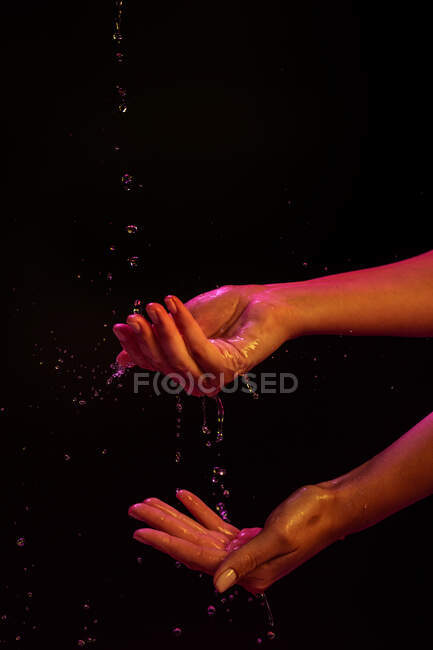 Vista Crop di donna anonima lavarsi le mani con spruzzi d'acqua sotto le luci al neon sullo sfondo nero — Foto stock