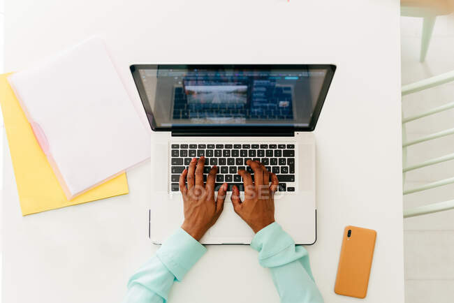 Vista dall'alto di ritaglio nero femminile digitando sulla tastiera del computer portatile mentre si lavora al tavolo bianco con documenti — Foto stock