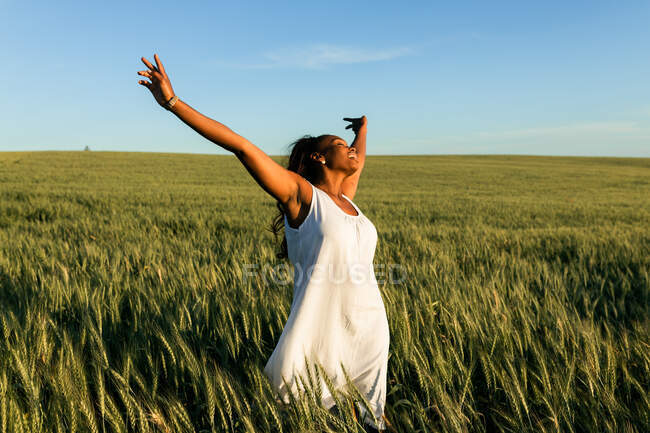 Sorrindo jovem senhora negra em vestido de verão branco passeando no campo de trigo verde, enquanto os olhos fechados durante o dia sob o céu azul — Fotografia de Stock