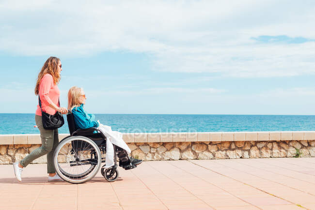 Side view of adult daughter walking with elderly mother in wheelchair along promenade near sea in summer — Stock Photo