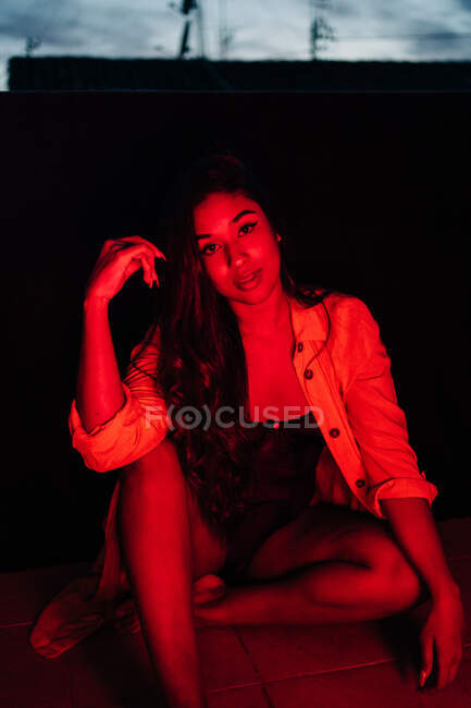 Sensual joven hispana étnica femenina en lencería mirando a la cámara mientras descansa en la terraza bajo la luz roja de neón por la noche - foto de stock