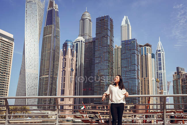 Low angle of happy young female tourist in casual outfit leaning back on railing and enjoying sunny day while standing on embankment against modern skyscrapers in Dubai Marina district of Dubai city — Stock Photo