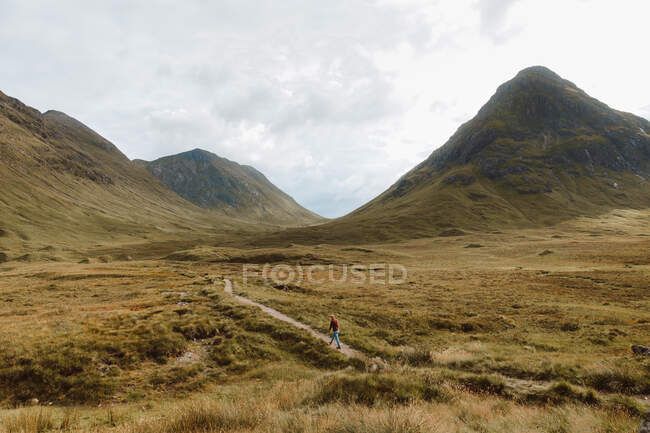 Vue arrière de l'homme méconnaissable marchant sur un chemin de terre sur une colline herbeuse rugueuse pendant le voyage à travers Glencoe dans la campagne britannique par temps nuageux — Photo de stock