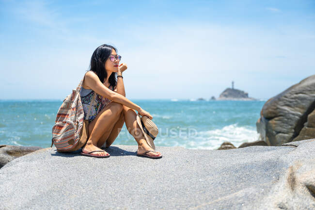 Schöne asiatische Mädchen sitzt auf einem Felsen und genießt das Meer an einem sonnigen Sommertag — Stockfoto