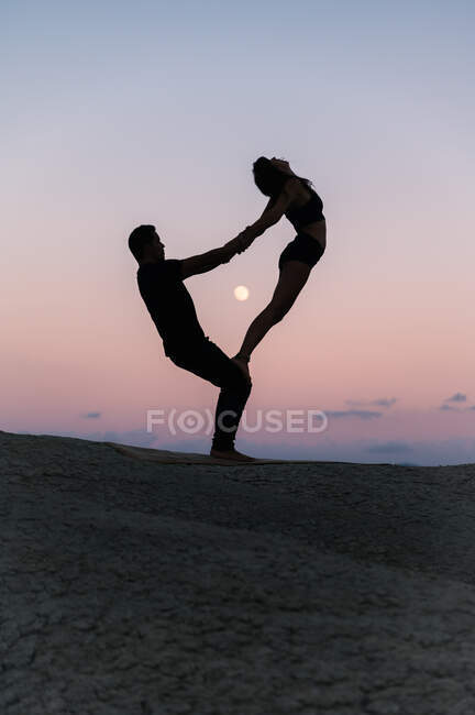 Vista laterale della silhouette di donna flessibile irriconoscibile in piedi sulle gambe dell'uomo durante la sessione di acro yoga sullo sfondo del cielo serale — Foto stock
