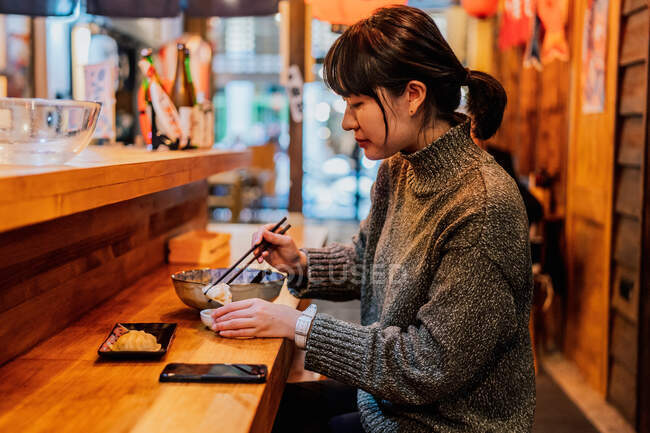 Vista laterale della donna asiatica soddisfatta in maglione sorridente mentre prende cucchiaio da lavoratore seduto al bancone di legno nel bar del ramen — Foto stock