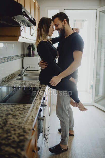 Allegro uomo barbuto che abbraccia donna sorridente mentre si appoggia su armadio con lavandino in cucina accogliente a casa — Foto stock