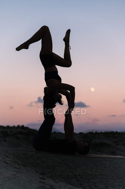 Vista lateral da mulher flexível equilibrando de cabeça para baixo enquanto pratica acroioga com parceiro masculino contra o céu do pôr-do-sol nas montanhas — Fotografia de Stock