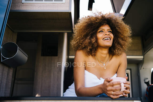 Low angle of relaxed African American female with mug of hot drink and chilling inside modern RV in summer morning — Stock Photo