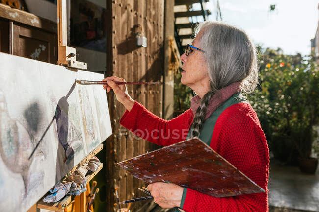 Side view of aged female artists in protective mask painting on canvas in backyard on sunny day in summer — Stock Photo