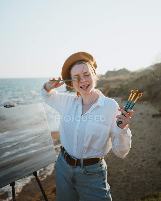 Young female artist in stylish outfit and beret standing with paintbrushes while grimacing with tongue out on shore near sand and sea while drawing painting on canvas on easel — Stock Photo