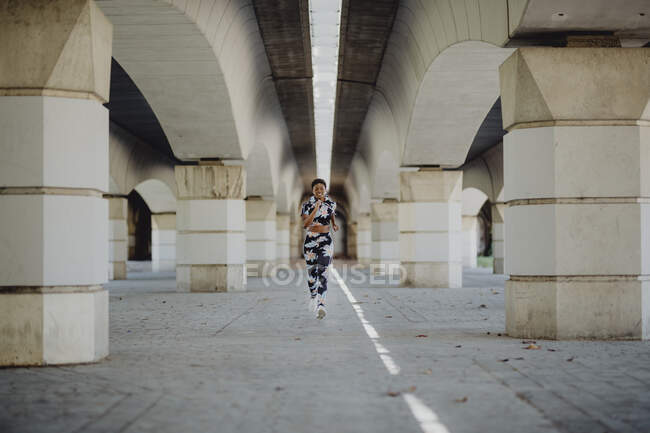 Young fitness African American woman running while looking at the camera on city street — Stock Photo