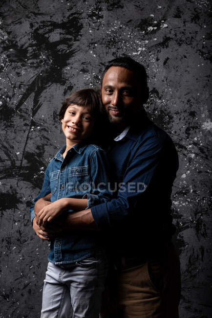 Веселий багаторасовий батько з сином-підлітком обіймають темне тло в студії і дивляться на камеру — стокове фото
