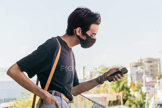 Vue latérale de l'homme homosexuel en masque de protection à l'aide d'un téléphone portable tout en se tenant sur le balcon en ville pendant le coronavirus — Photo de stock