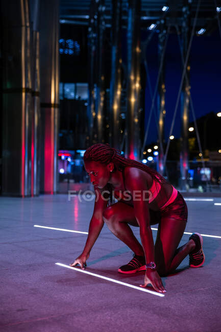 Vista lateral de la mujer atlética étnica en agacharse inicio preparado para el sprint en la calle por la noche - foto de stock