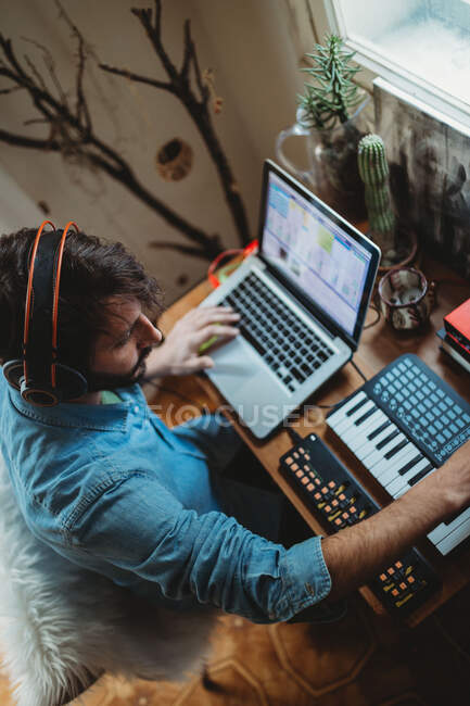 From above side view of focused young man in headphones working on synthesizer and laptop at table at home — Stock Photo