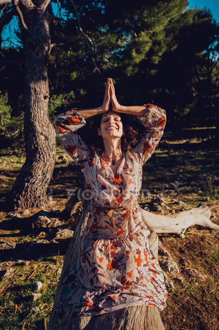Cheerful female in dress practicing yoga meditation on Hero pose while sitting on tree trunk in highlands on sunny day with yes closed — Stock Photo