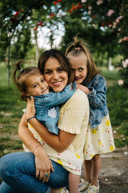 Jovem mãe feliz com filhinhas bonitos em roupas semelhantes desfrutando de dia ensolarado de verão juntos enquanto sentados no parque — Fotografia de Stock