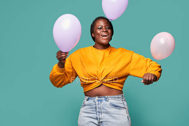 Femme afro-américaine excitée en jeans et haut jaune debout près de ballons tombants sur fond bleu en studio — Photo de stock