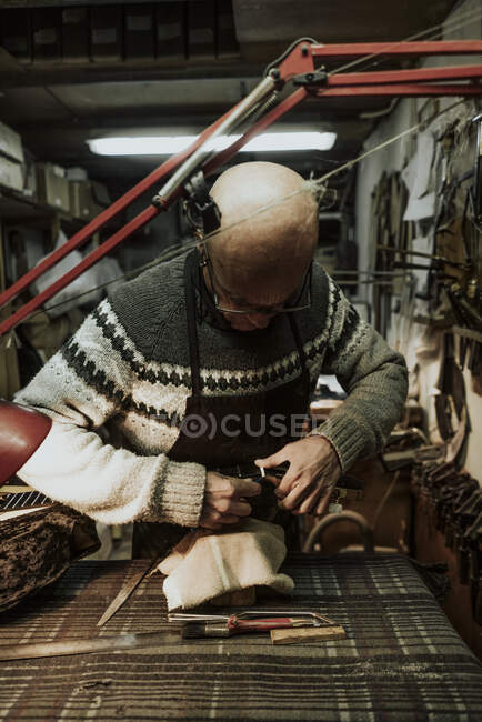 Концентрированный пожилой лысый ремесленник в повседневной теплой одежде и очках, тюнинг струн акустической гитары во время работы в старомодной мастерской — стоковое фото