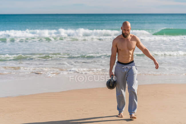 Hombre deportivo sin camisa llevando kettlebell caminando por la costa arenosa con olas del océano en el fondo mirando hacia abajo - foto de stock