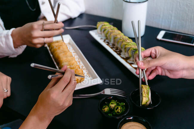 High angle of crop anonymous friends with reusable chopsticks eating delicious sushi rolls with soy sauce and jalapeno pepper — Stock Photo