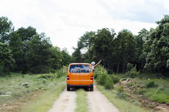 Безтурботні анонімні мандрівники з витягнутими руками, що керують помаранчевим фургоном вздовж дороги в лісі під час літньої подорожі — стокове фото