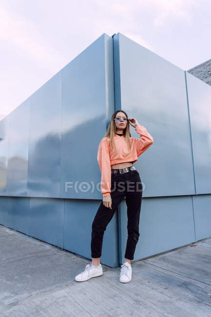 Уверенная женщина в джинсах с верхней и оранжевой рубашкой со стильными солнцезащитными очками, стоящими у стены — стоковое фото