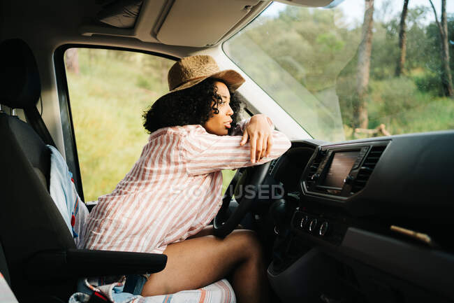 Vista lateral de la joven viajera afroamericana romántica reflexiva sentada en el asiento del conductor y mirando hacia otro lado mientras disfruta del viaje de verano en la naturaleza - foto de stock