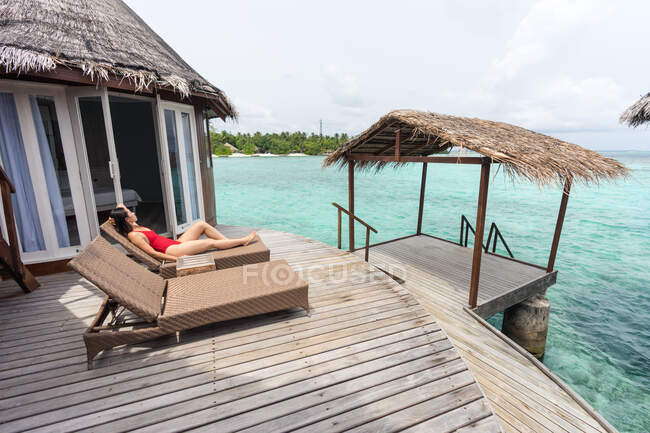 Vista lateral da fêmea com a mão atrás da cabeça em maiô deitado na cadeira de praia relaxante em Maldivas — Fotografia de Stock