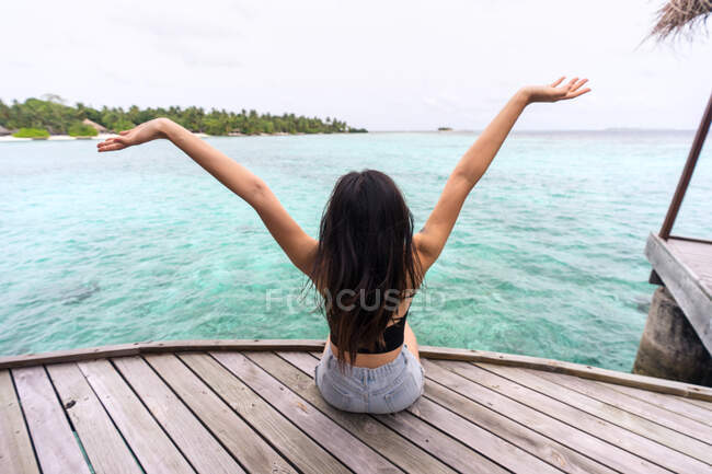 Rückansicht einer nicht wiedererkennbaren Frau in lässiger Kleidung, die auf einem Holzsteg sitzt und es sich auf den Malediven gemütlich macht — Stockfoto