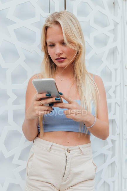 Mujer tranquila con el pelo rubio y en traje de verano de pie en la ciudad de navegación en el teléfono móvil - foto de stock