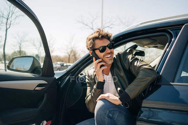 Happy male in sunglasses talking on cellphone while sitting in automobile on sunny day — Stock Photo