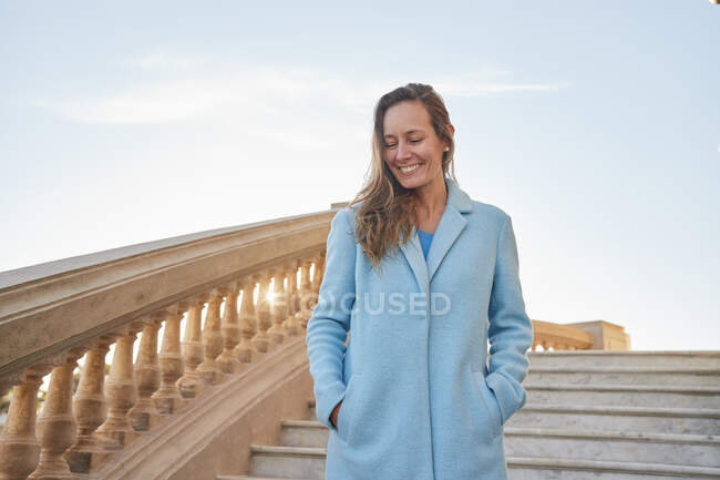 Niedriger Winkel einer positiven erwachsenen Frau in blauem Mantel, die an sonnigen Tagen auf Steinstufen mit Steingeländer unter blauem Himmel in der Stadtstraße spaziert — Stockfoto