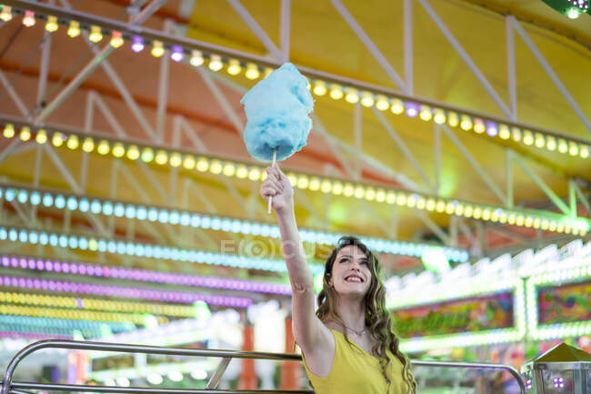 Optimistic female standing with blue cotton candy in raised arm while enjoying weekend in amusement park and looking up — Stock Photo