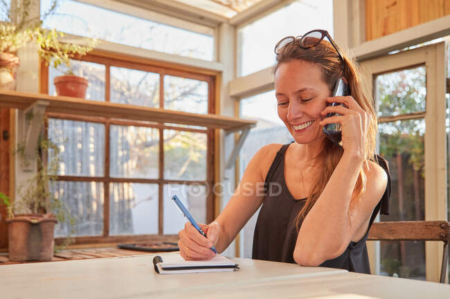 Усміхнена жінка садівник говорить на мобільному телефоні і робить нотатки, сидячи за столом і працюючи в теплиці в саду — стокове фото