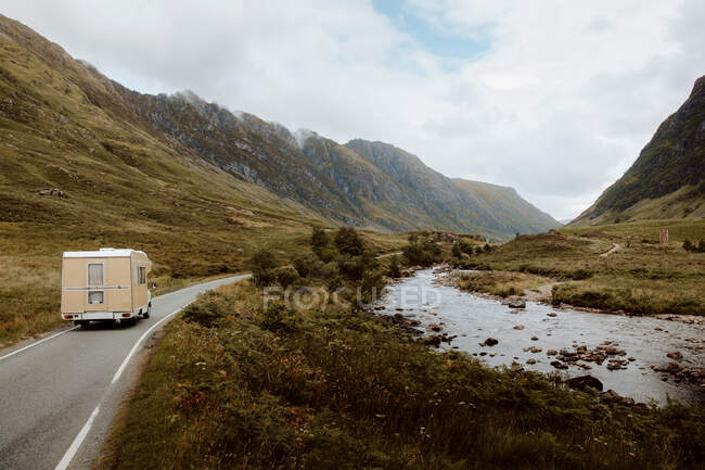 Caravan riding along asphalt road near brook amidst hills during trip through UK countryside on cloudy day — Stock Photo