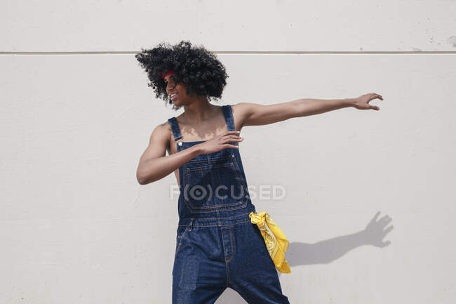 Cheerful African American male with Afro hairstyle in denim overalls looking away while dancing on concrete wall — Stock Photo