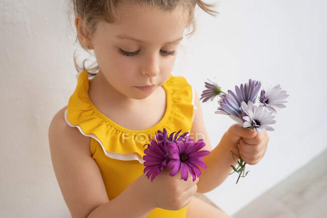 Charming thoughtful little girl in bright yellow swimsuit with blooming flowers in hands against white wall — Stock Photo