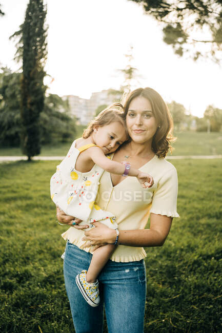 Sonriente madre joven llevando linda hija pequeña en vestido de verano con pulsera y mirando a la cámara mientras está de pie en el césped verde en el parque de verano - foto de stock