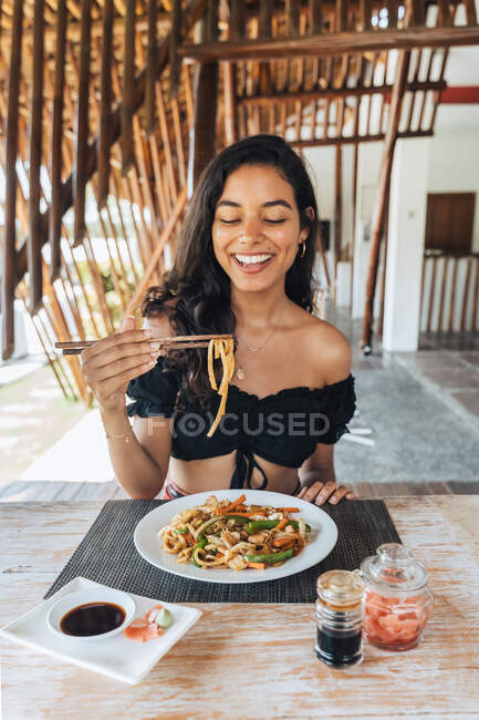 Touriste féminine joyeuse avec de délicieuses pâtes entre les bâtonnets de nourriture au-dessus de la table avec de la sauce soja et des tranches de gingembre marinées à l'extérieur — Photo de stock