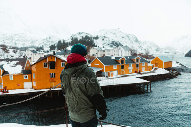 Vista posterior del hombre anónimo en ropa de abrigo parado en la orilla del mar contra las casas amarillas y la cresta de montaña nevada en el día de invierno en las Islas Lofoten, Noruega - foto de stock