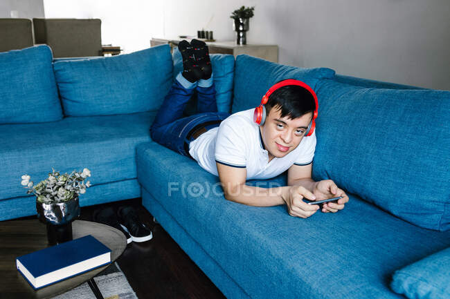 Deleitado adolescente latino en auriculares jugando videojuego en el teléfono móvil mientras está acostado en el sofá en casa y mirando a la cámara - foto de stock