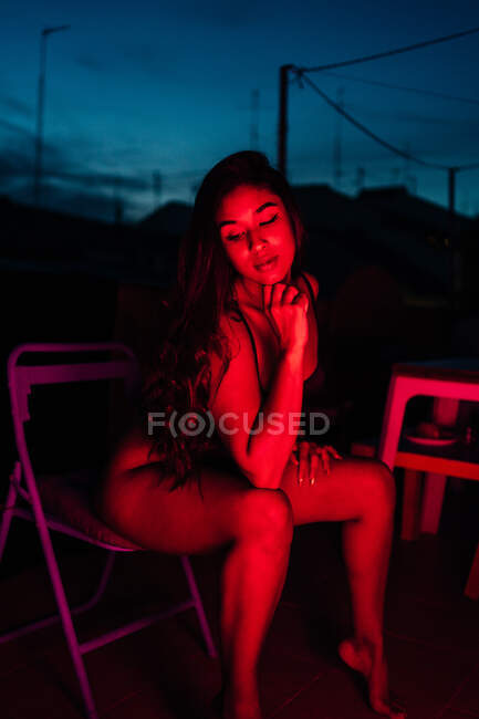 Молода жінка в нижній білизні дивиться вниз, сидячи на стільці під червоним неоновим світлом вночі на терасі — стокове фото
